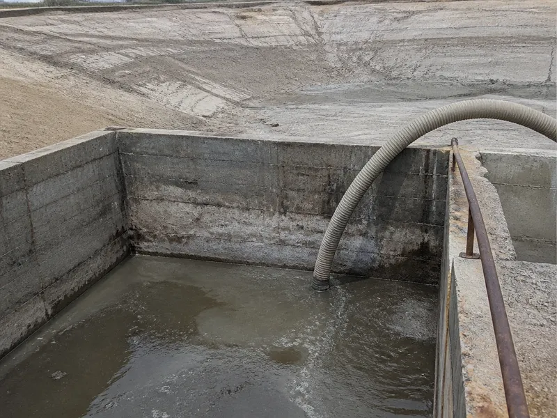 Foso industrial con lodo y agua y manguera de aspiración, con el talud de una balsa de riego al fondo